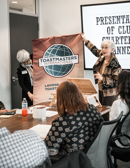 Nancy Ducket and Hallie Adams unveil the LAVIDGE Ad Libs Toastmasters banner at a charter and officer installation ceremony Feb. 5, 2020.